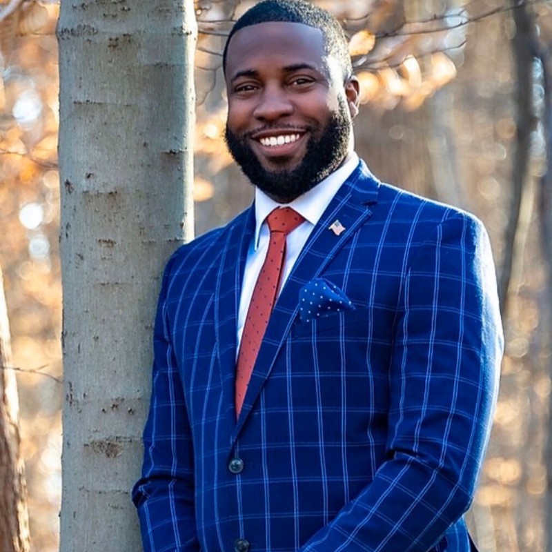 Photo of a man om a blue suit with a red tie smiling and standing next to a tree